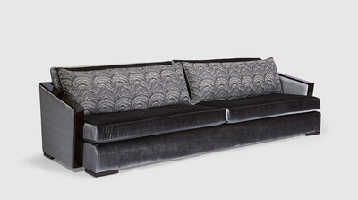 An art deco inspired sofa that is upholstered in a gun metal grey velvet which is accented by two, long scatter cushions. It also features diagonal arms which are enhanced by a dark mahogany detail, front view.