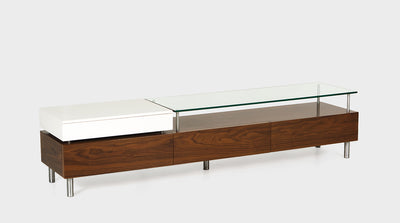 A contemporary TV unit with three walnut drawers and one contrasting, white drawer. It has a glass top and aluminum legs. 