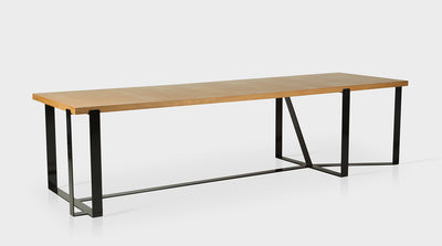 A contemporary dining table with a rectangular, oak top and a black, powder coated, steel base that has an unconventional geometric design.