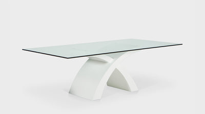 A contemporary dining table with a clear, glass, rectangular top and a white, timber base made up of two intersecting crescents. 