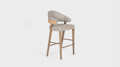 A contemporary barstool that has a grey, upholstered seat with a rounded back and arms. It has a double fabric detail on the back in a tan coloured fabric and it has diagonal, oak legs.