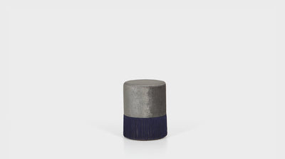 A modern ottoman that is fully upholstered in a silver grey fabric with a royal blue tassel detail.