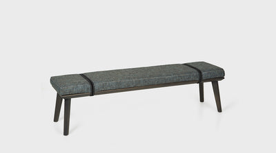 A contemporary ottoman that has a blue grey upholstered seat with a black, wraparound, strap detail. It has a solid oak base with diagonal legs. 