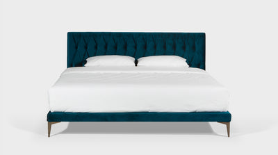 A fully upholstered bed with a rich, blue, diamond button head board and timber legs, front view.